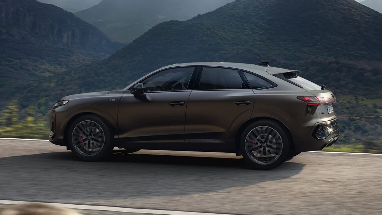 Ein brauner Audi Q3 Sportback fährt auf einer kurvigen Straße durch eine bergige Landschaft. Im Hintergrund sind bewaldete Hügel und ein bewölkter Himmel zu sehen. 