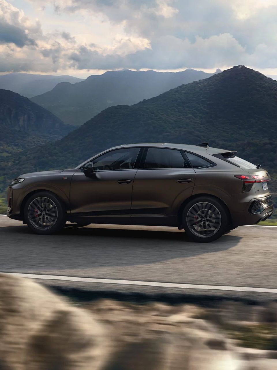 Ein brauner Audi Q3 Sportback fährt auf einer kurvigen Straße durch eine bergige Landschaft. Im Hintergrund sind bewaldete Hügel und ein bewölkter Himmel zu sehen. 