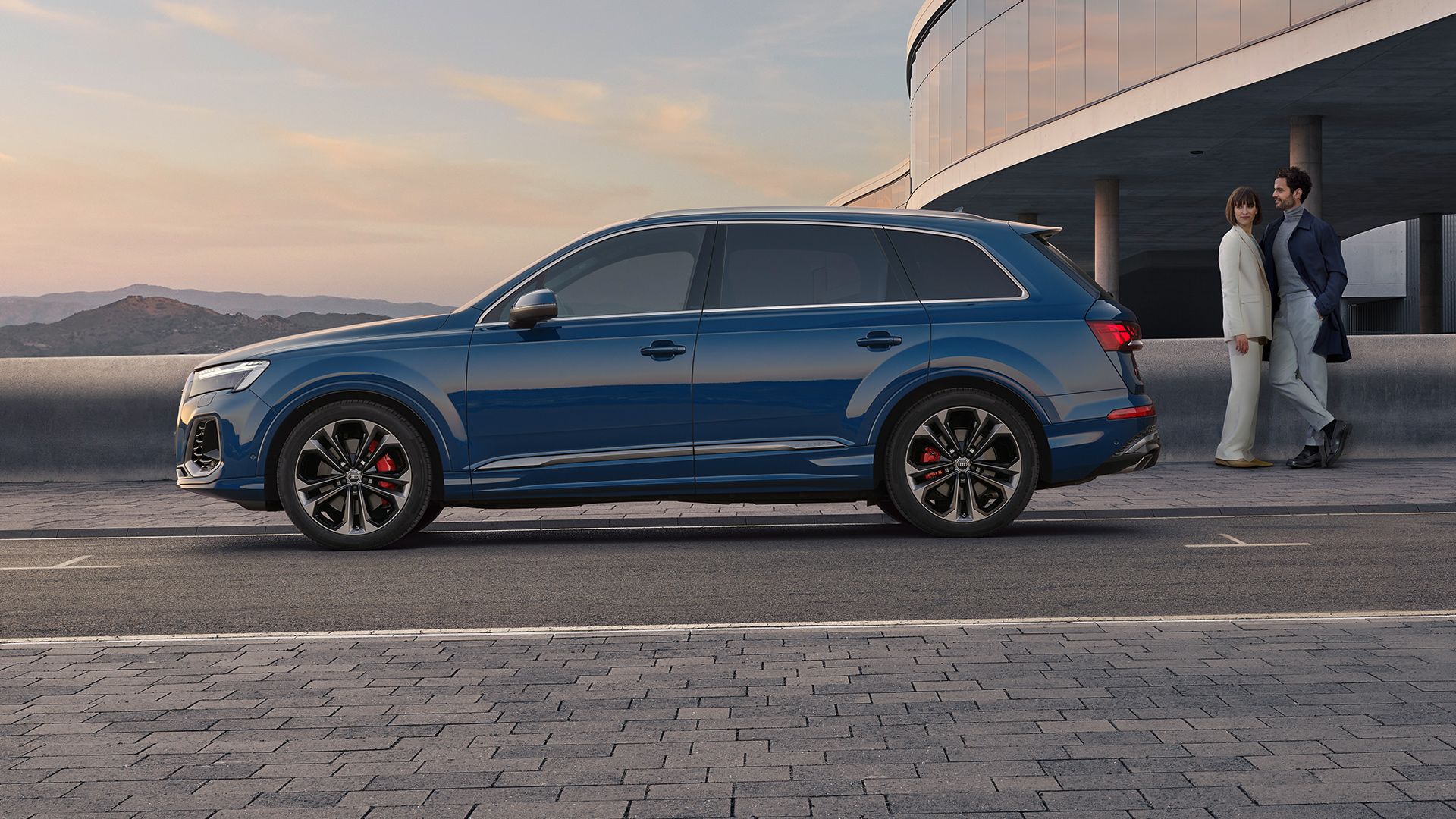 Син Q7 SUV автомобил е паркиран пред модерна, извита сграда. Двама души, облечени в формални костюми, вървят по тротоара до автомобила. Небето е ясно, с лека облачност при залез слънце.