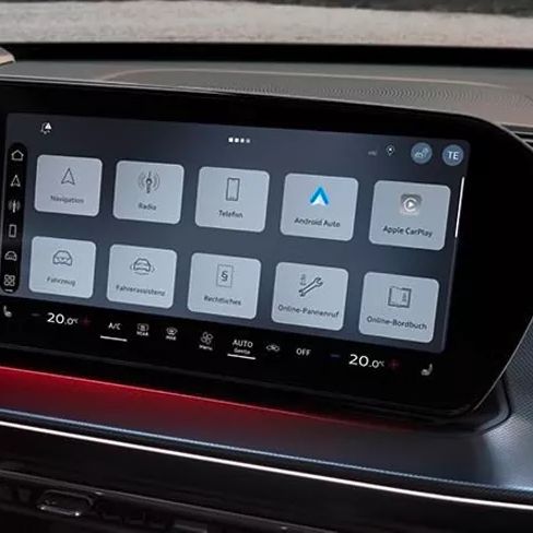 Das Bild zeigt den Innenraum eines Audi Q3 SUV und einen großen Bildschirm auf der Mittelkonsole. Auf dem Bildschirm sind verschiedene Bedienungsoptionen für Navigation, Radio, Smartphone-Integration und Fahrzeugfunktionen sichtbar. Links vom Bildschirm ist teilweise das Lenkrad zu sehen.