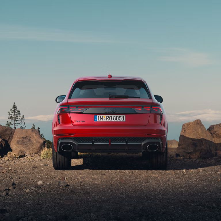 Der RS Q8 SUV performance von hinten parkend auf einem Schotterweg. Links und rechts sind kleine Felsen erkennbar, dahinter einige Bäume und ein Wolkenband.