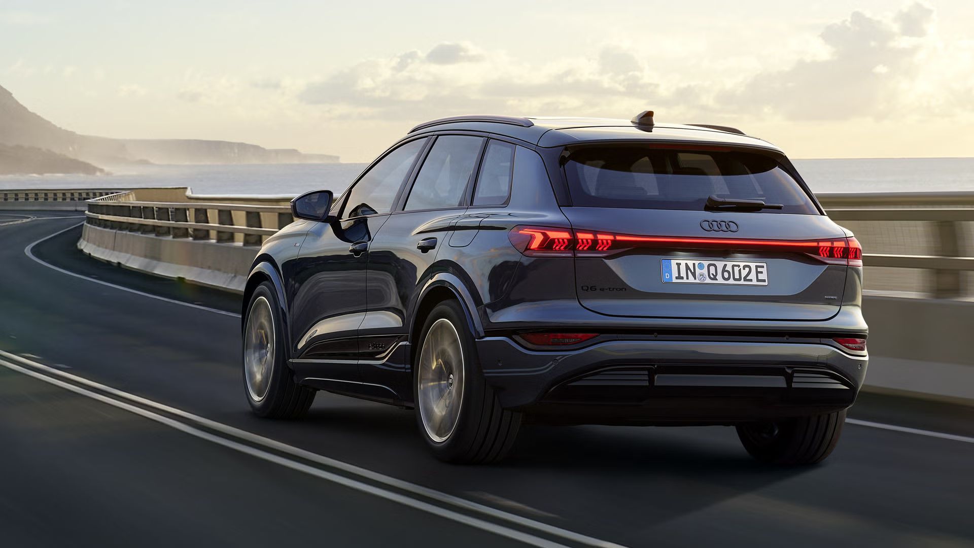  Q6 SUV e-tron отзад, който се движи по наклонен път край морето. На заден план се виждат планини.