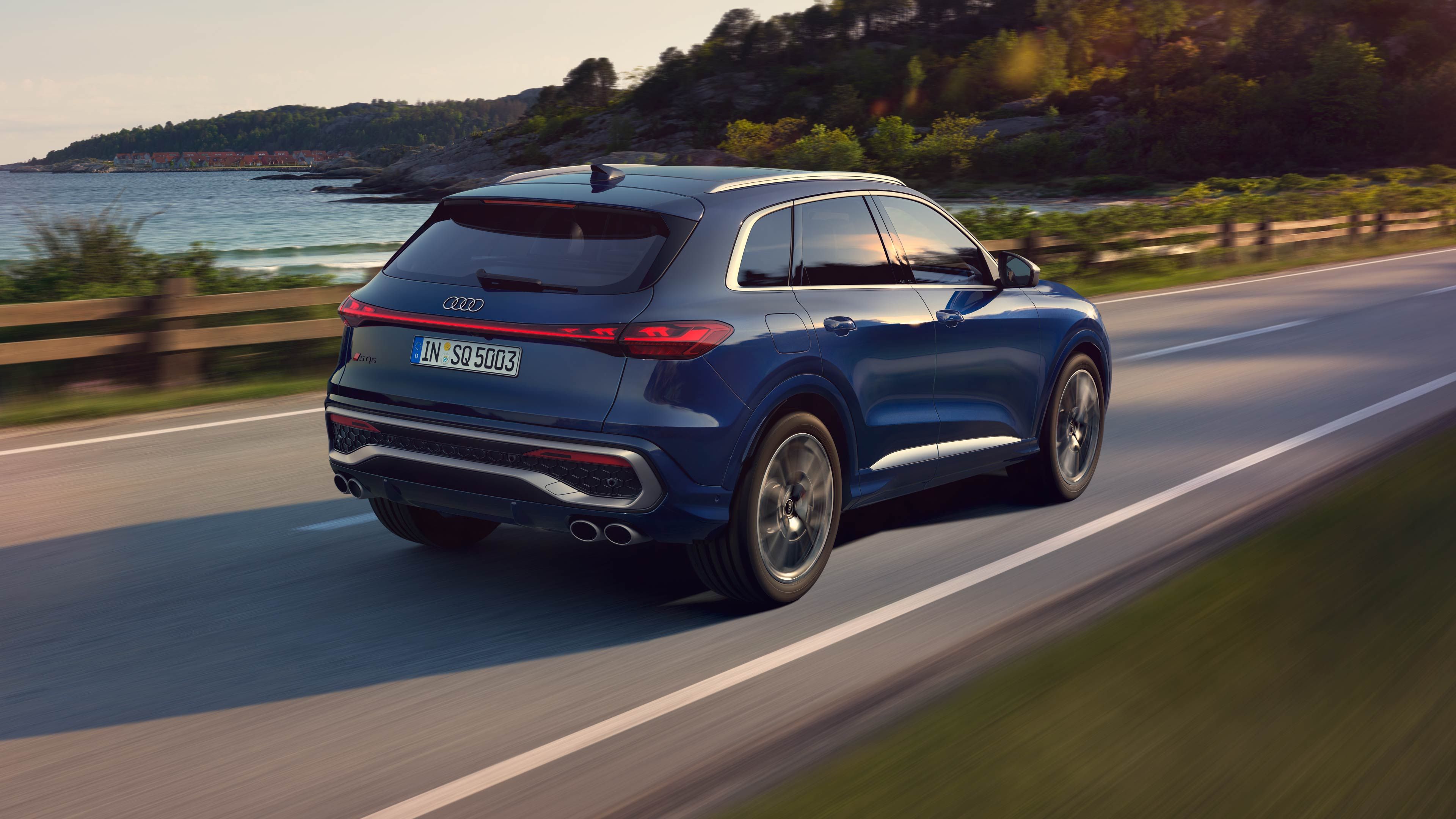 Син Audi SQ5 на път край гора и езеро през деня. Колата е заснета отзад и малко настрани, като се вижда регистрационния номер. Областта около пътя е зелена и живописна.