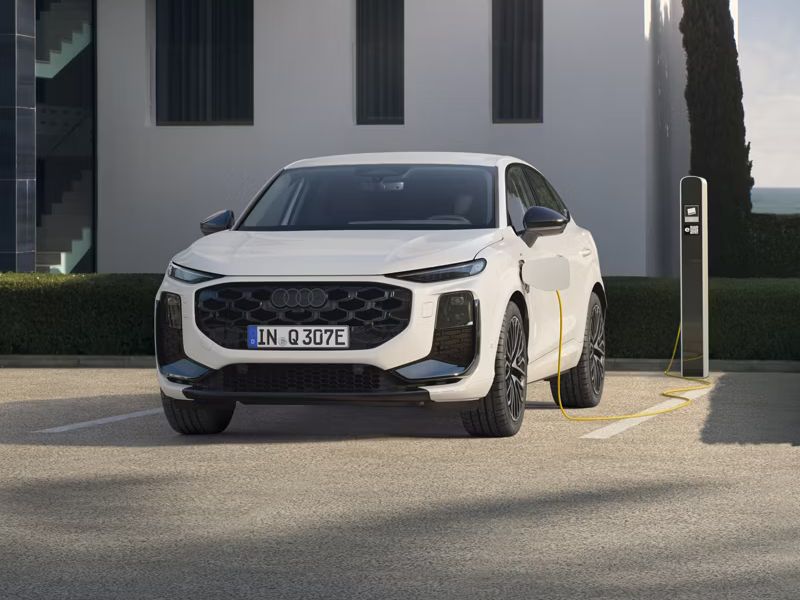 Auf dem Bild ist der Audi Q3 Sportback e-hybrid zu sehen. Dieser steht vor einem Wohngebäude mit einer Ladesäule und lädt.