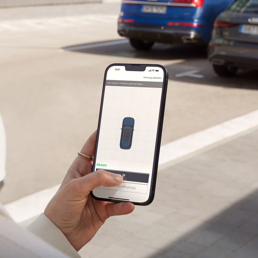 Eine modisch gekleidete junge Frau hält ein Smartphone in der Hand, auf dem ein stilisierter Audi in Draufsicht zu erkennen ist. Sie steht vor parkenden Fahrzeugen.