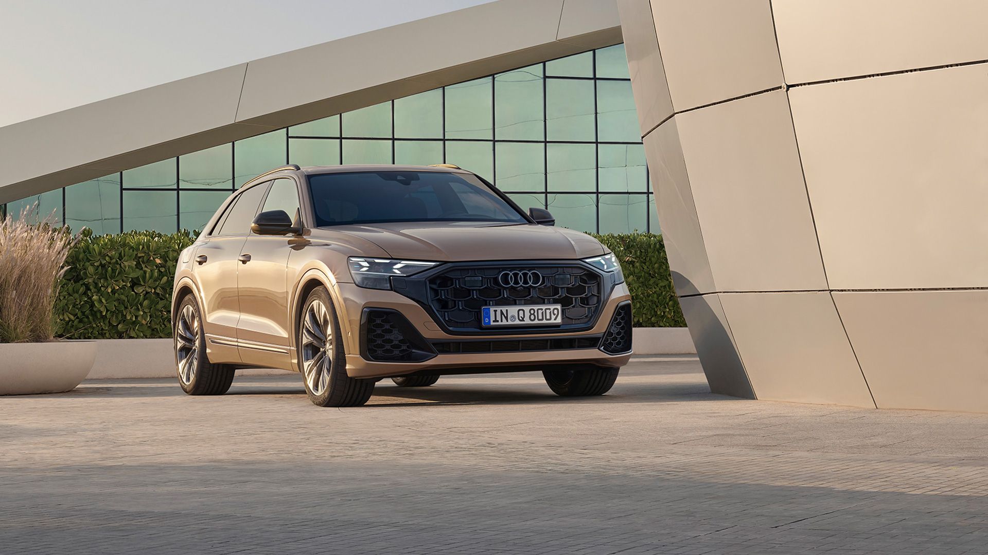 Луксозен кафяво-бронзов автомобил Audi Q8 е паркиран пред модерна сграда с геометрични форми. На заден план има зеленина и прозорци с огледални стъкла. Колата изглежда стилна и съвременна.